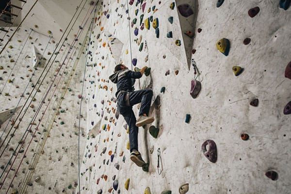A climber in dark clothing ascends a large indoor climbing wall with various colorful handholds.