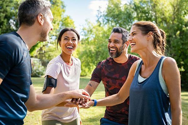 Four people in activewear smiling and placing their hands together in a huddle outdoors on a sunny day.