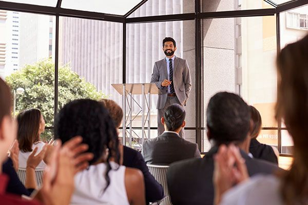 A presenter in a grey suit stands at a podium in a sunlit conference room as audience members applaud.