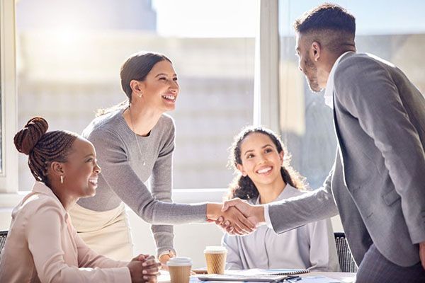 Four colleagues in an office setting smiling as two of them shake hands over a table with coffee cups.