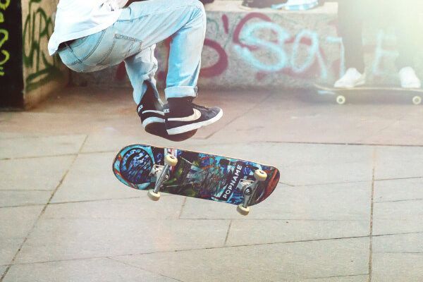 A skateboarder performs a trick in mid-air above a paved ground with graffiti-covered structures in the background.