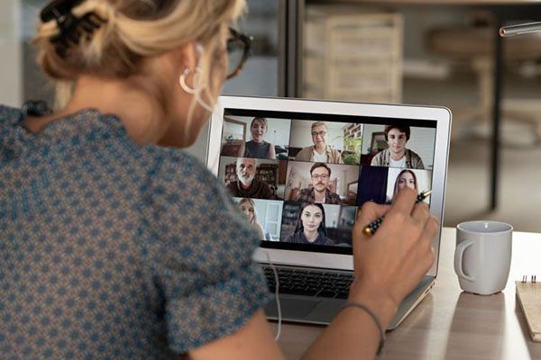 A person in a blue patterned shirt uses a laptop for a video conference with multiple participants on screen.