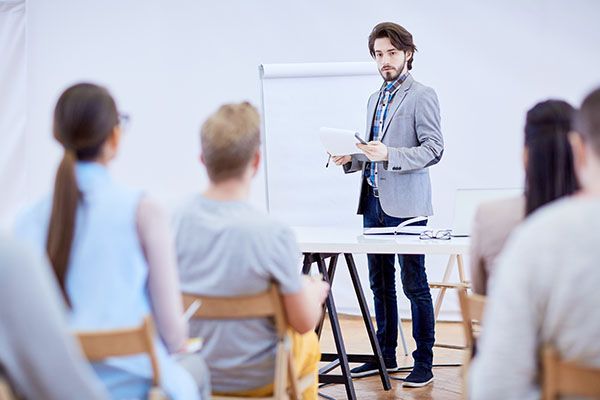 A person in a gray blazer presents to a group of students sitting in a bright, modern classroom.