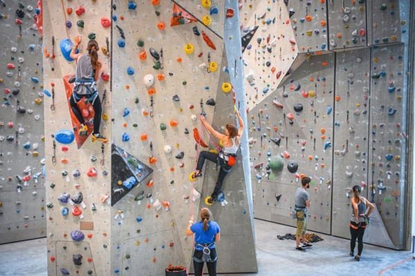 Four people in a climbing gym: two scaling rock walls with holds of various colors and two standing on the floor below.