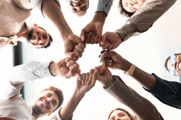 A diverse group of people join their fists together in a circle, viewed from below against a bright, clear background.