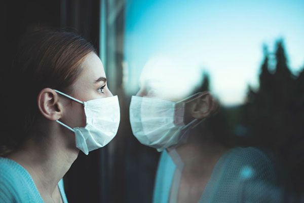 A person wearing a white face mask looking at their reflection in a window.