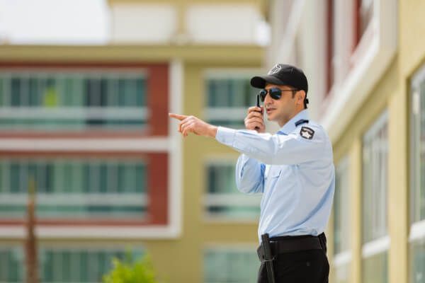 A security guard in a light blue uniform and sunglasses stands outside, talking on a radio and pointing to the side.