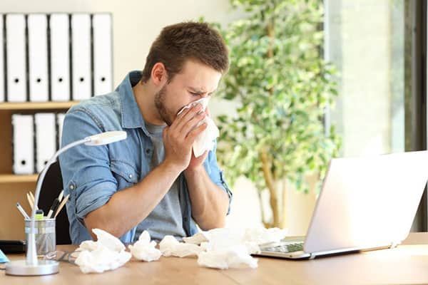 A person sitting at a desk with a laptop blows their nose into a tissue, surrounded by used tissues in an office setting.