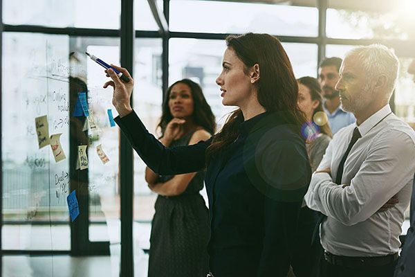 A group of professional colleagues brainstorm at a glass wall covered in sticky notes in an office setting.
