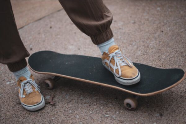 A person in brown joggers and light blue socks wearing yellow sneakers, one foot placed on a black skateboard.