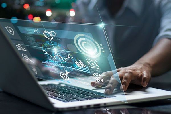 A person uses a laptop displaying a futuristic, glowing digital interface with data charts and icons in a dark room.