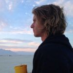 A profile view of a person with wavy hair looking out over a wide, pale landscape under a soft-colored sky.