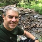 Man with graying hair smiles, sitting beside a rocky stream in a forest.