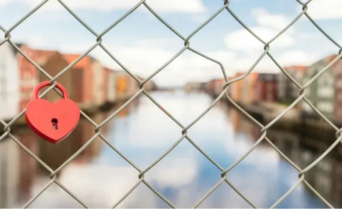 Red heart-shaped lock on a chain-link fence, blurred cityscape and river in the background.
