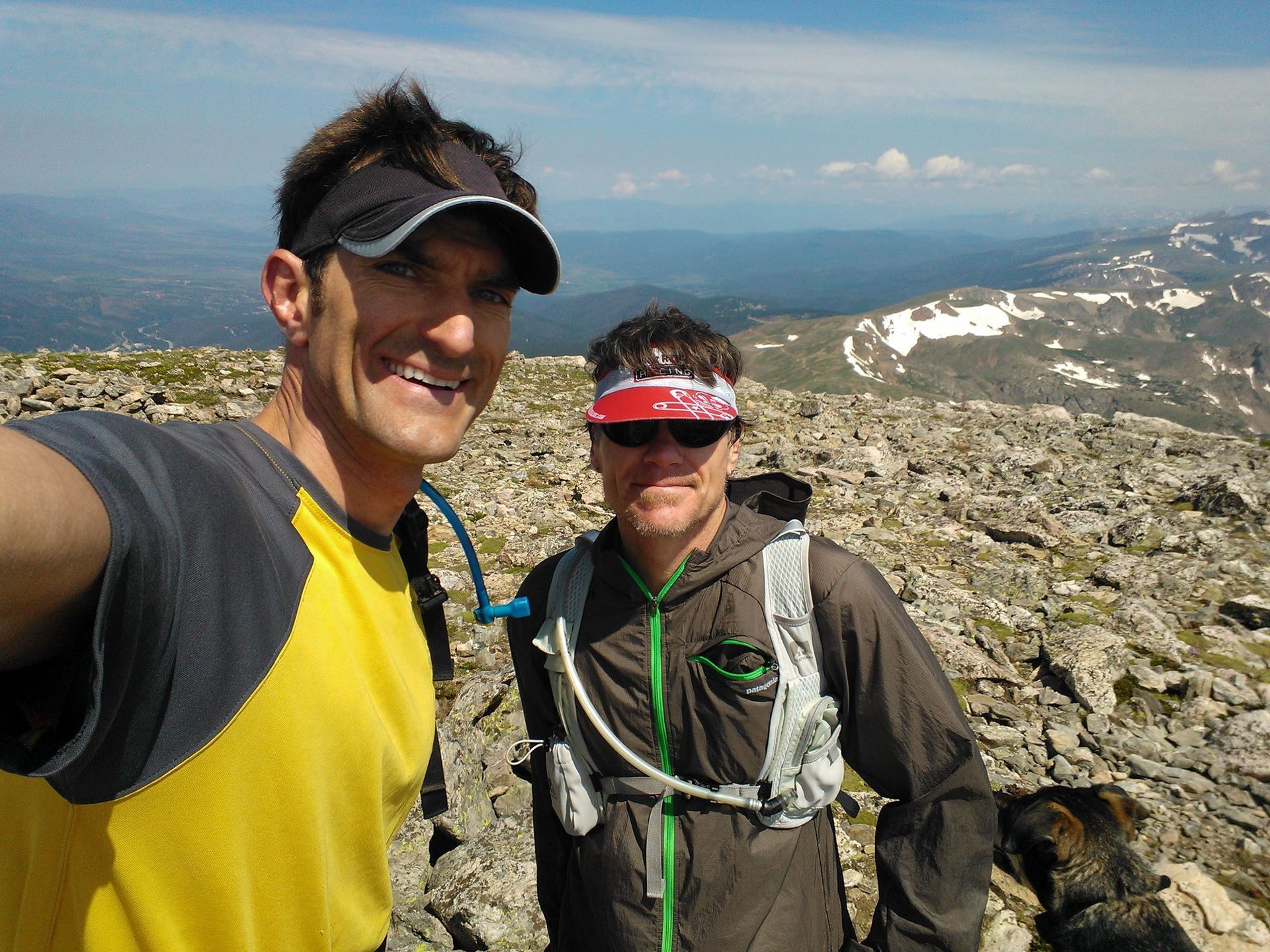 Two hikers taking a selfie on a rocky mountain summit; sunny day, blue sky; dog on right.