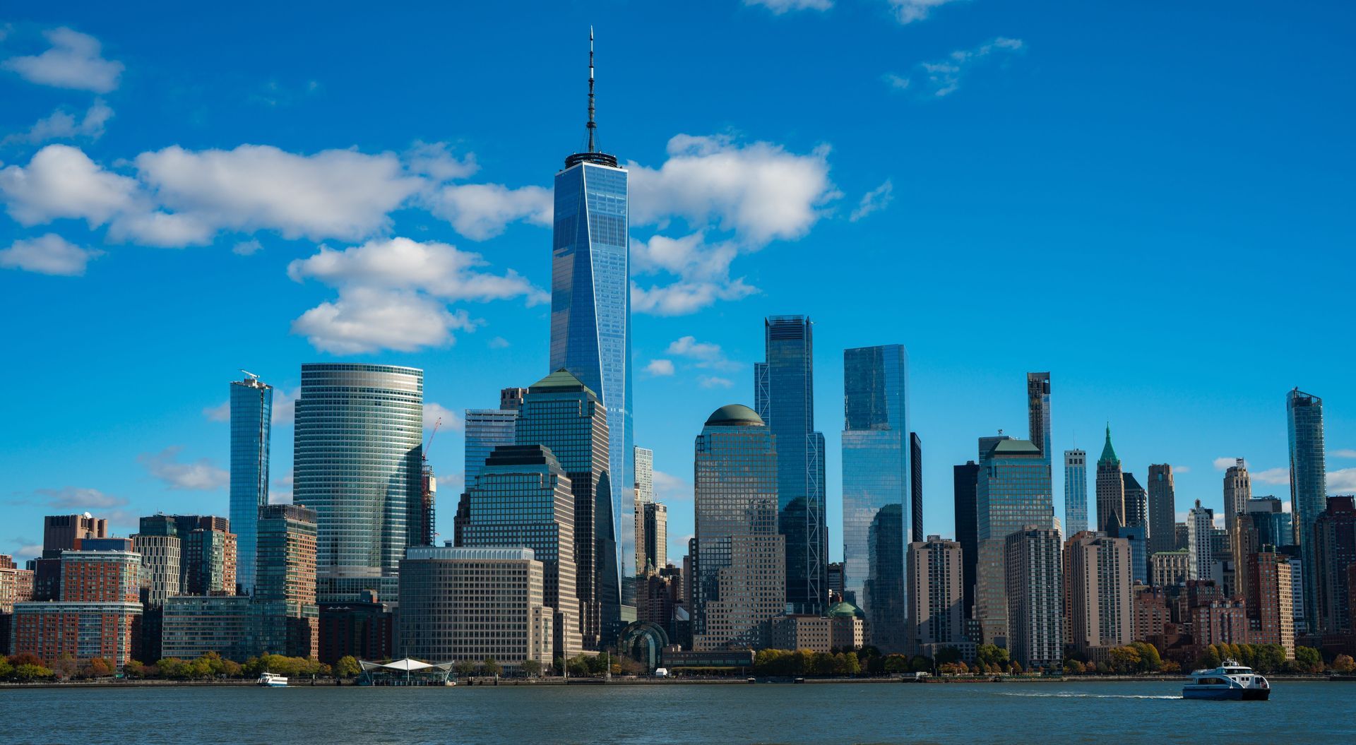 New York City skyline on a bright, sunny day with a blue sky and water in the foreground.