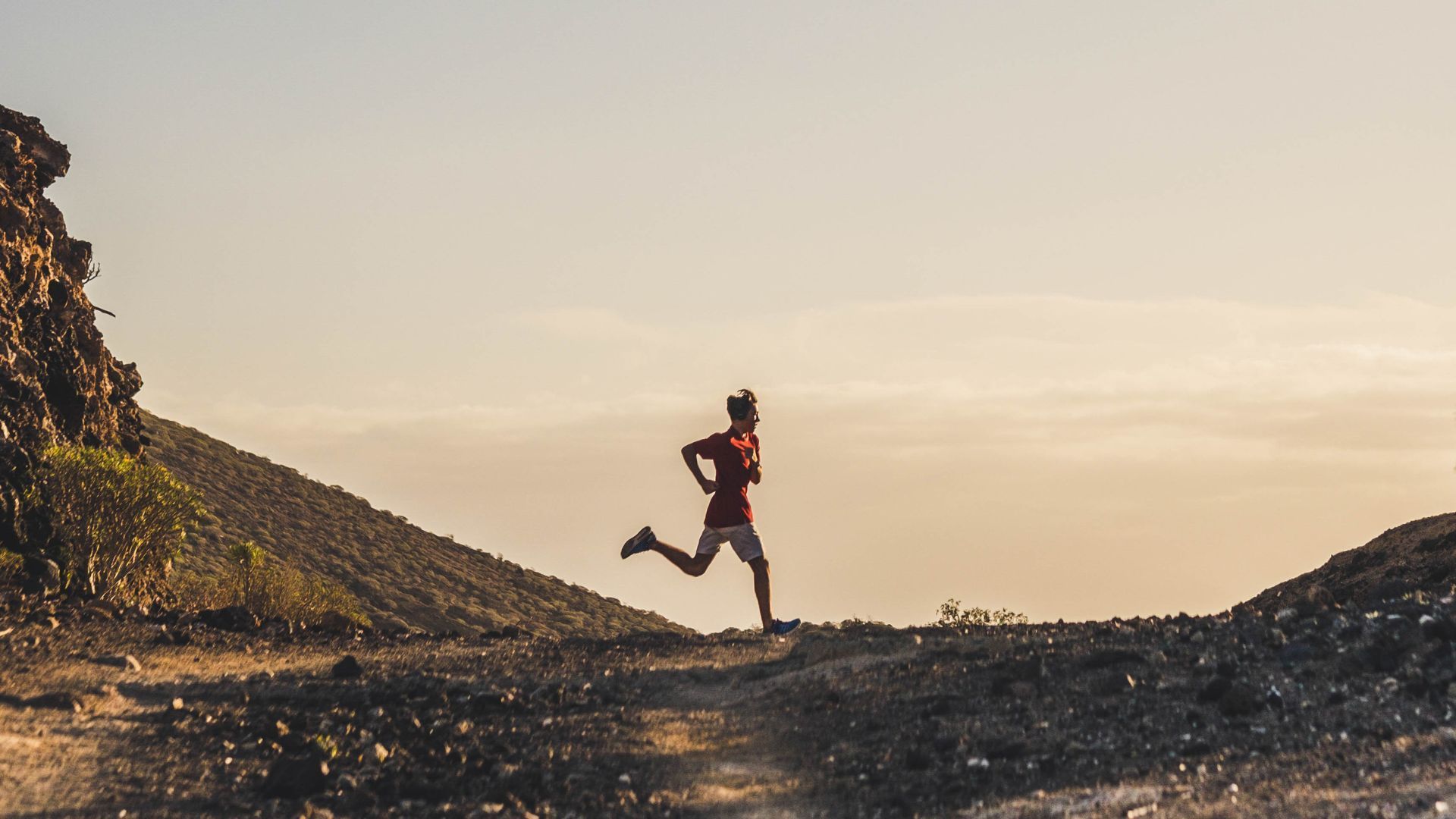 Runner silhouetted at sunset on a rocky trail, mid-stride between hills.