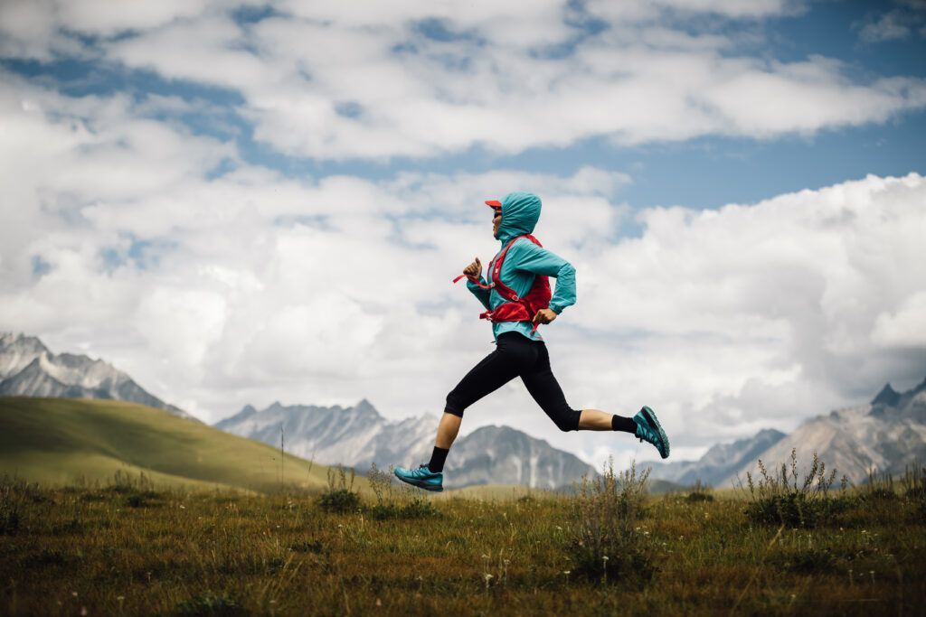 Runner in blue and red gear sprints across grassy terrain, mountains in background, cloudy sky.