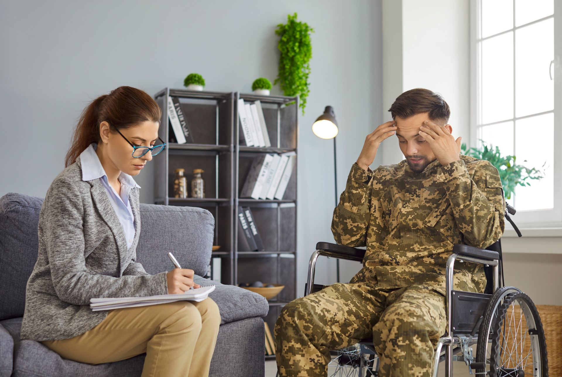 A therapist writes on a notepad while a soldier in uniform sits in a wheelchair, holding their head in distress.