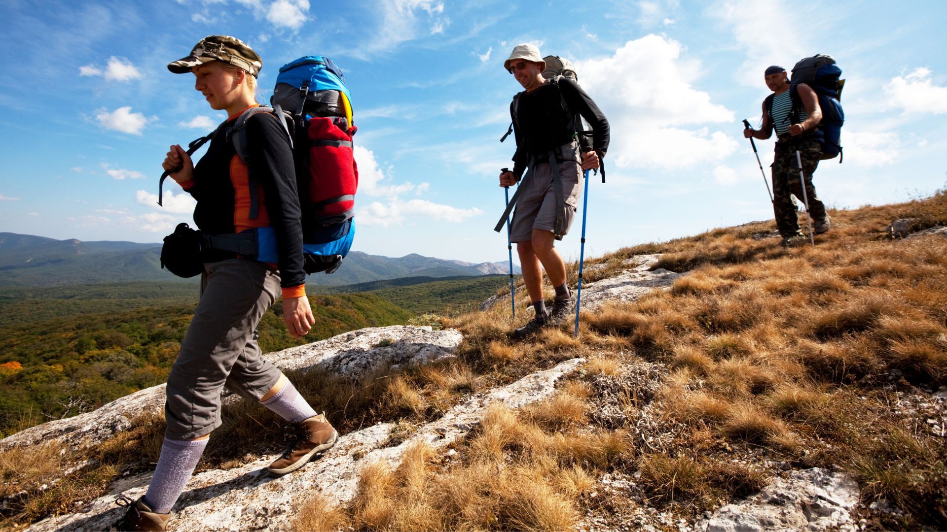 Four hikers with backpacks walking along a rocky mountain ridge under a blue sky