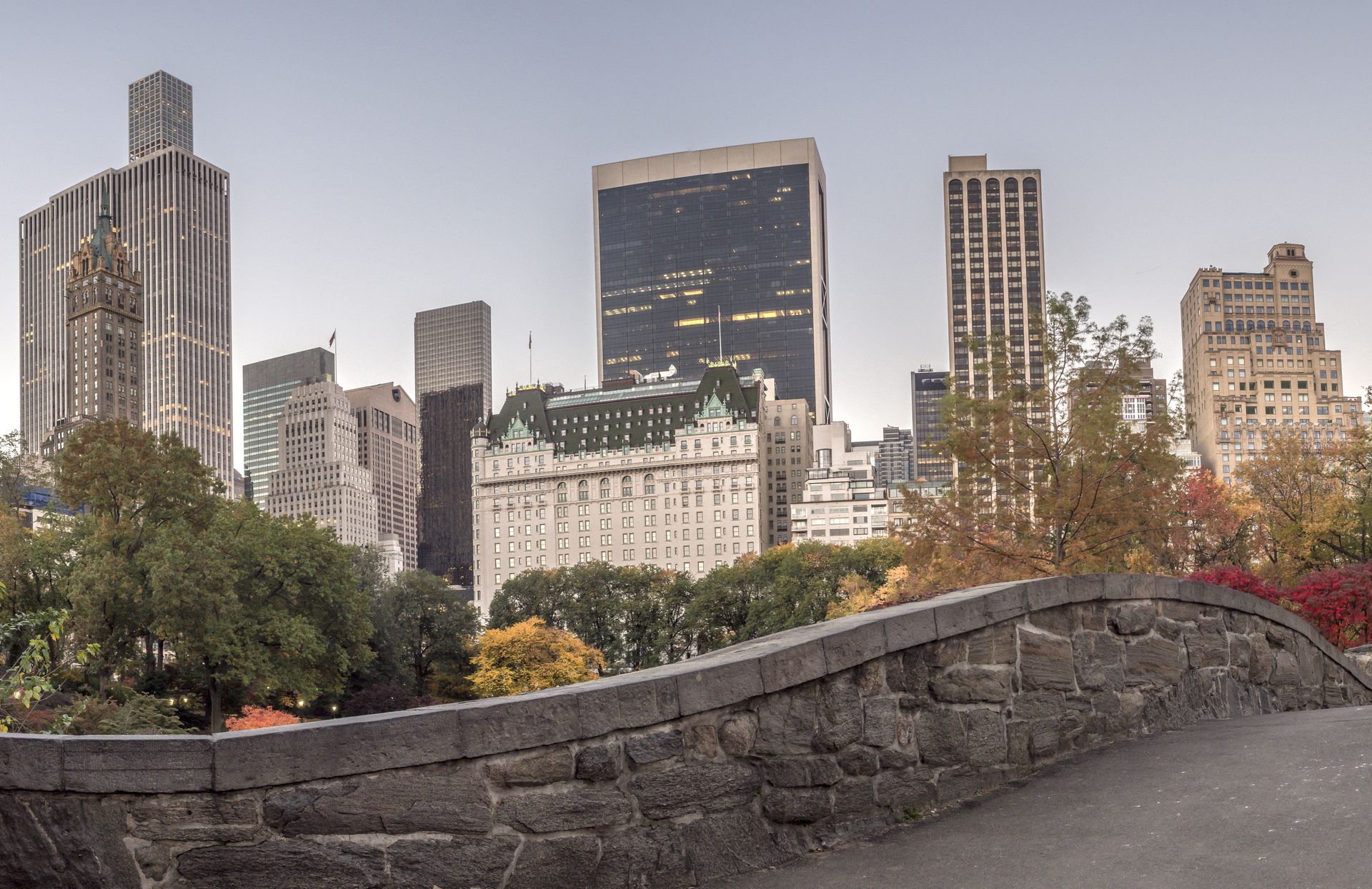 Skyline of New York City, including several skyscrapers, viewed from a stone bridge in a park.