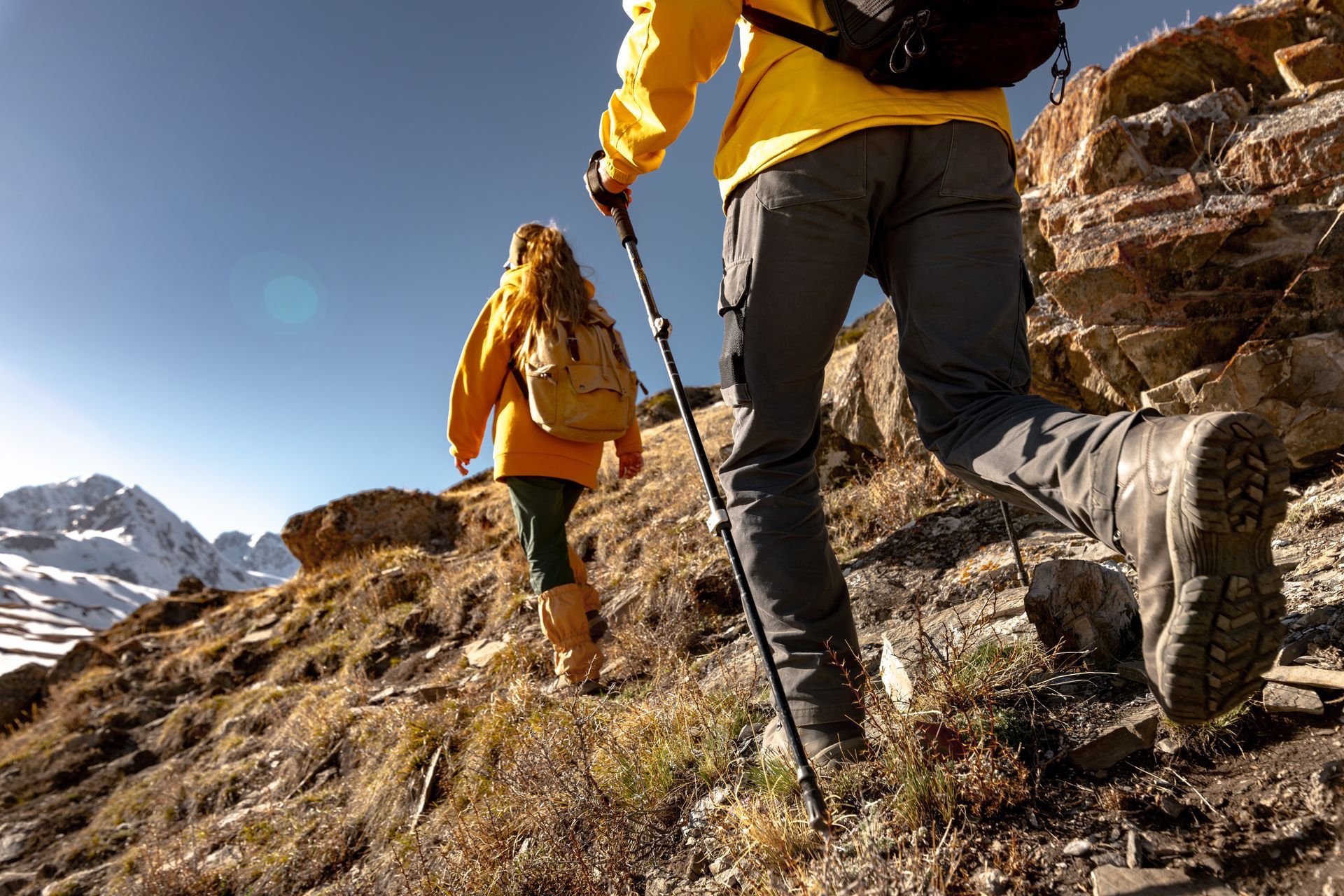 Two hikers in yellow jackets walk up a rocky mountain trail, with one using a trekking pole in the foreground.