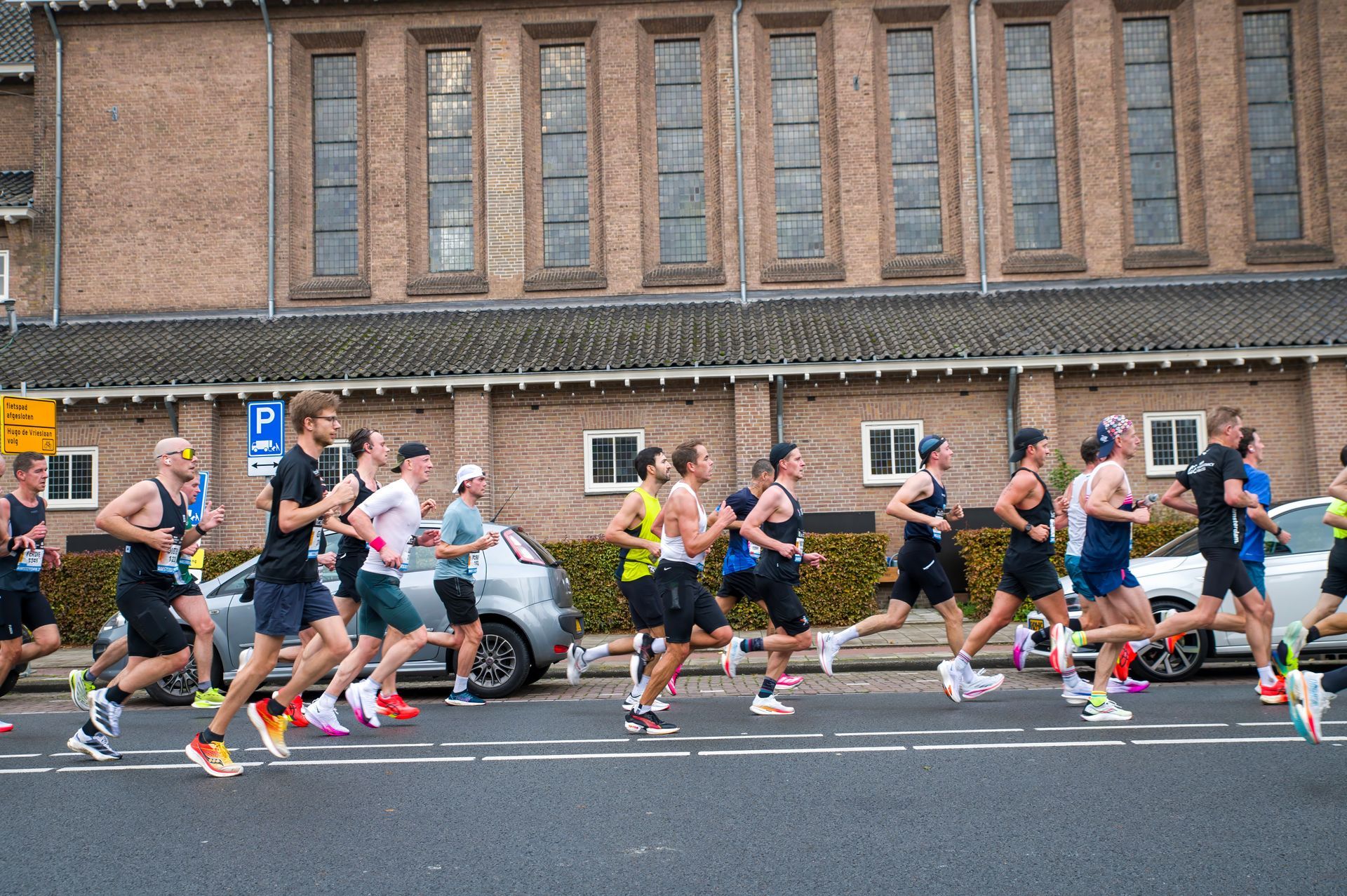 A group of runners in athletic gear race along a road in front of a brick building with arched windows.