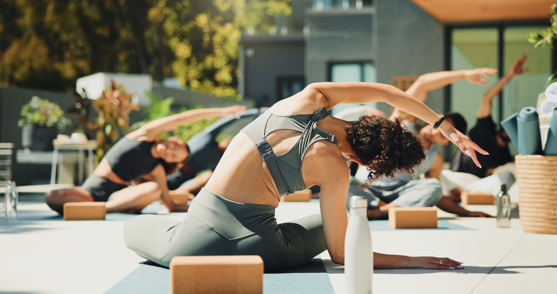 A group of people in athletic wear practices yoga poses on mats outdoors, stretching their arms overhead.