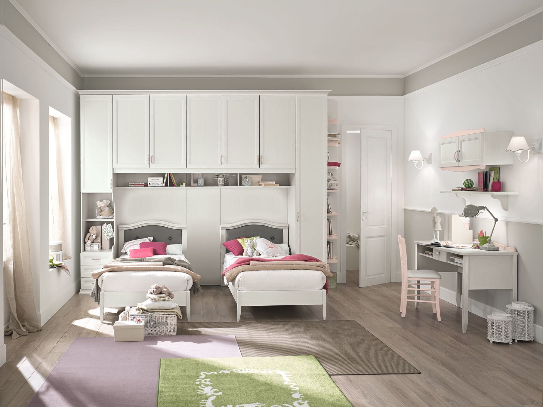 Una camera da letto luminosa con due letti, una scrivania e pensili. Dominano i colori bianco e pastello.