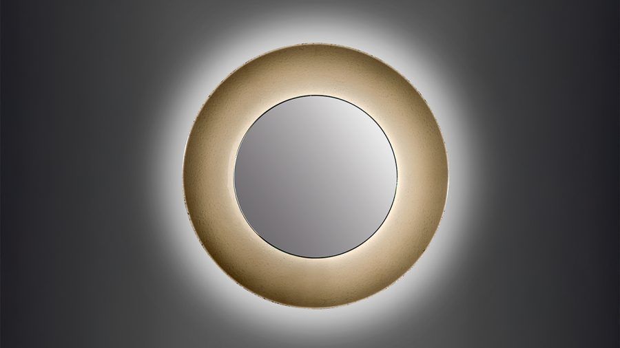 Specchio circolare con bordo dorato, retroilluminato con una luce bianca e soffusa su una parete grigia.