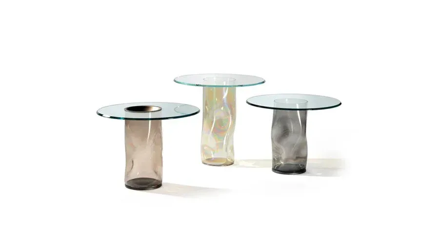 Tre tavolini laterali con piano in vetro e basi colorate e testurizzate nei colori marrone chiaro, crema e grigio.