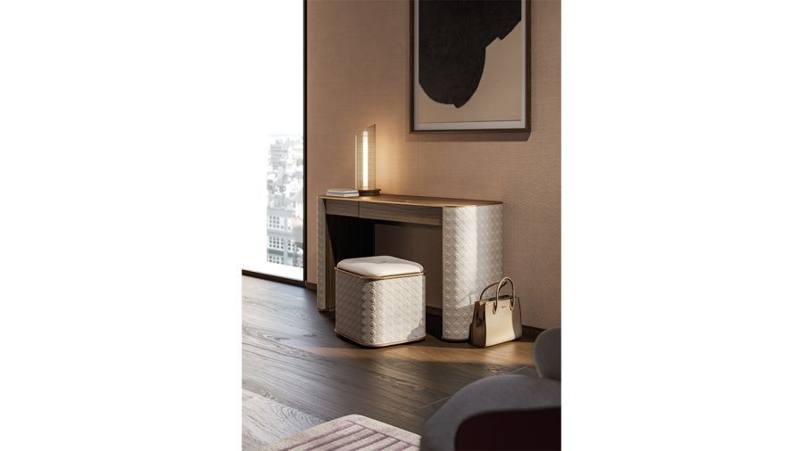 Una scrivania, uno sgabello e una lampada in una stanza con opere d'arte, una borsa e una finestra. I toni neutri dominano la scena.