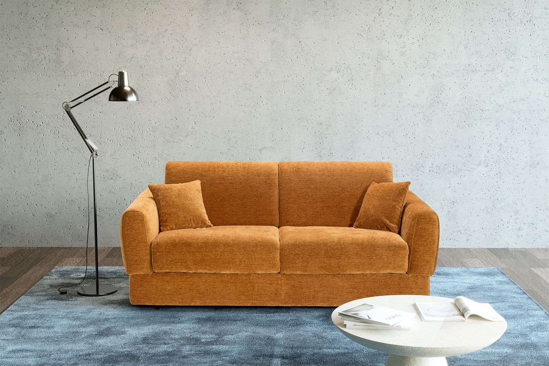 Divano arancione con cuscini, lampada da terra e tavolino da caffè su un tappeto blu, contro un muro di cemento.