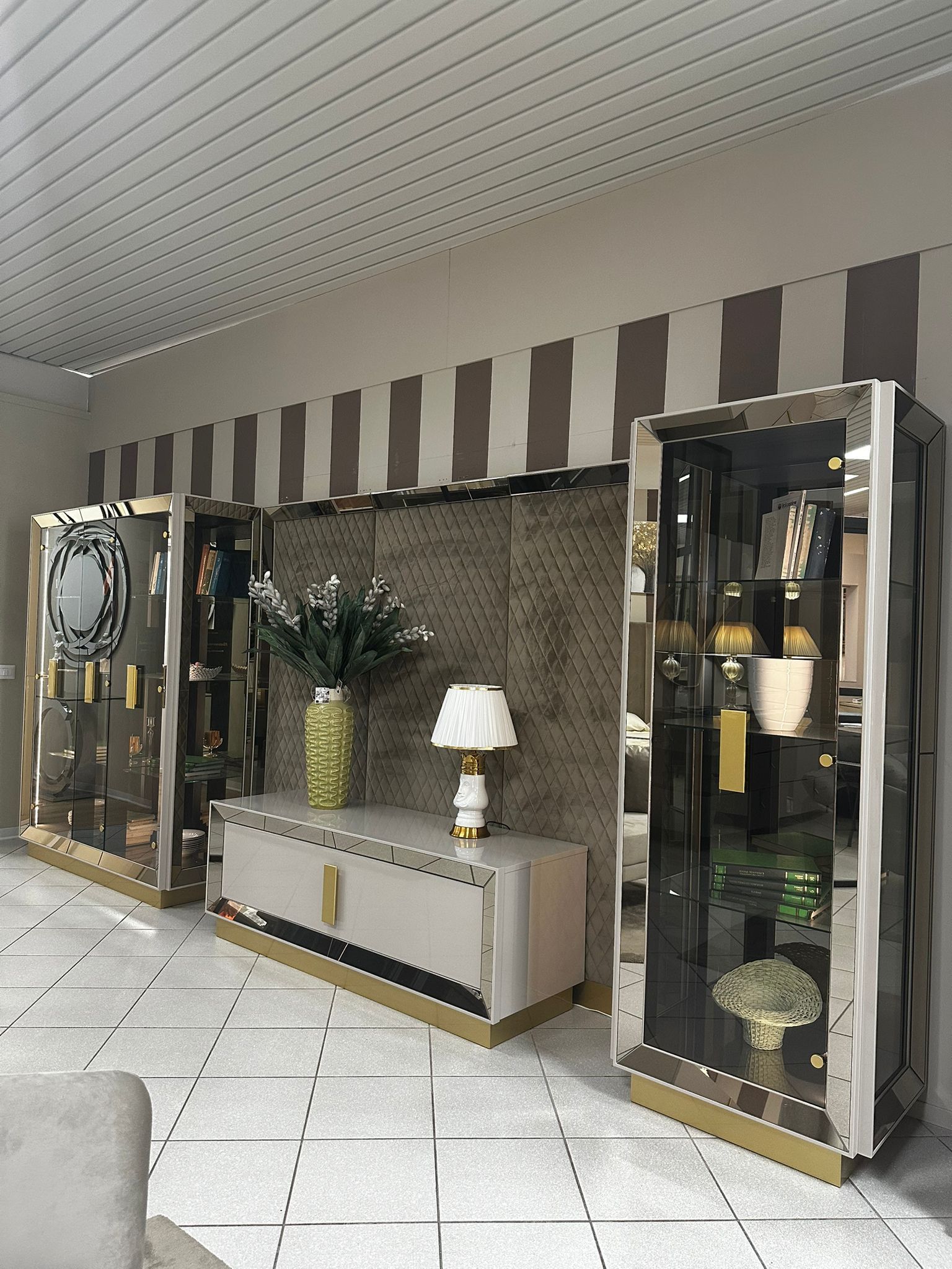 Elegante set di mobili: vetrine con finiture dorate, una consolle e uno sfondo a righe in un interno ben illuminato.