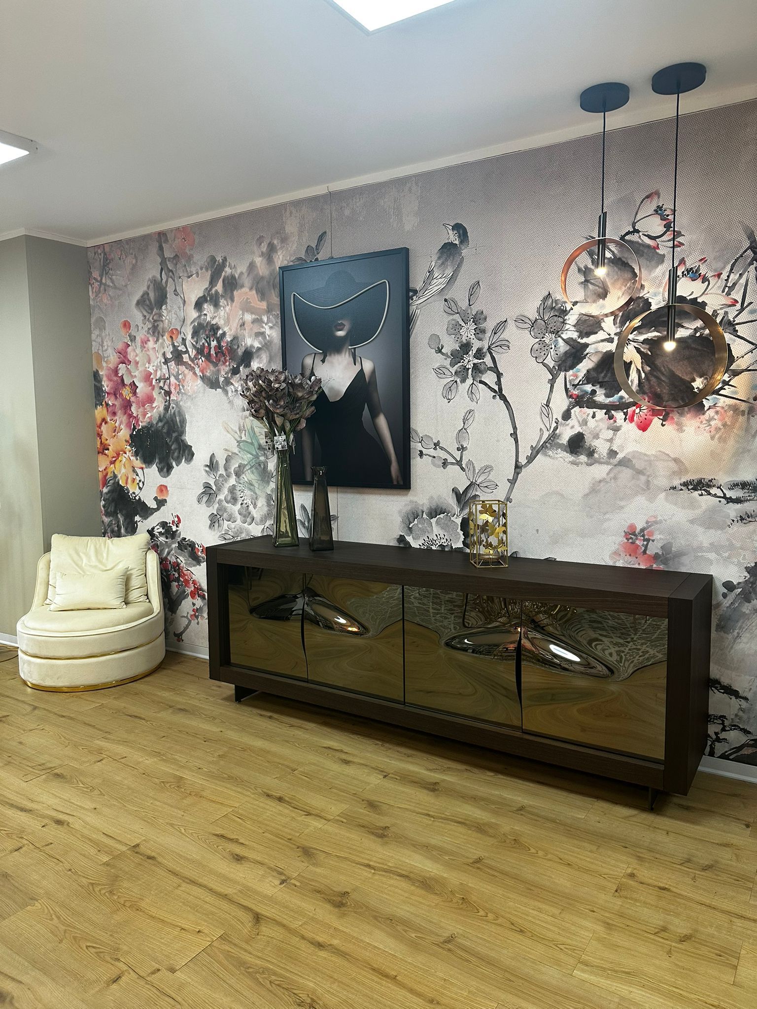 Un soggiorno con carta da parati floreale, credenza in legno scuro, quadri neri e sedia bianca.