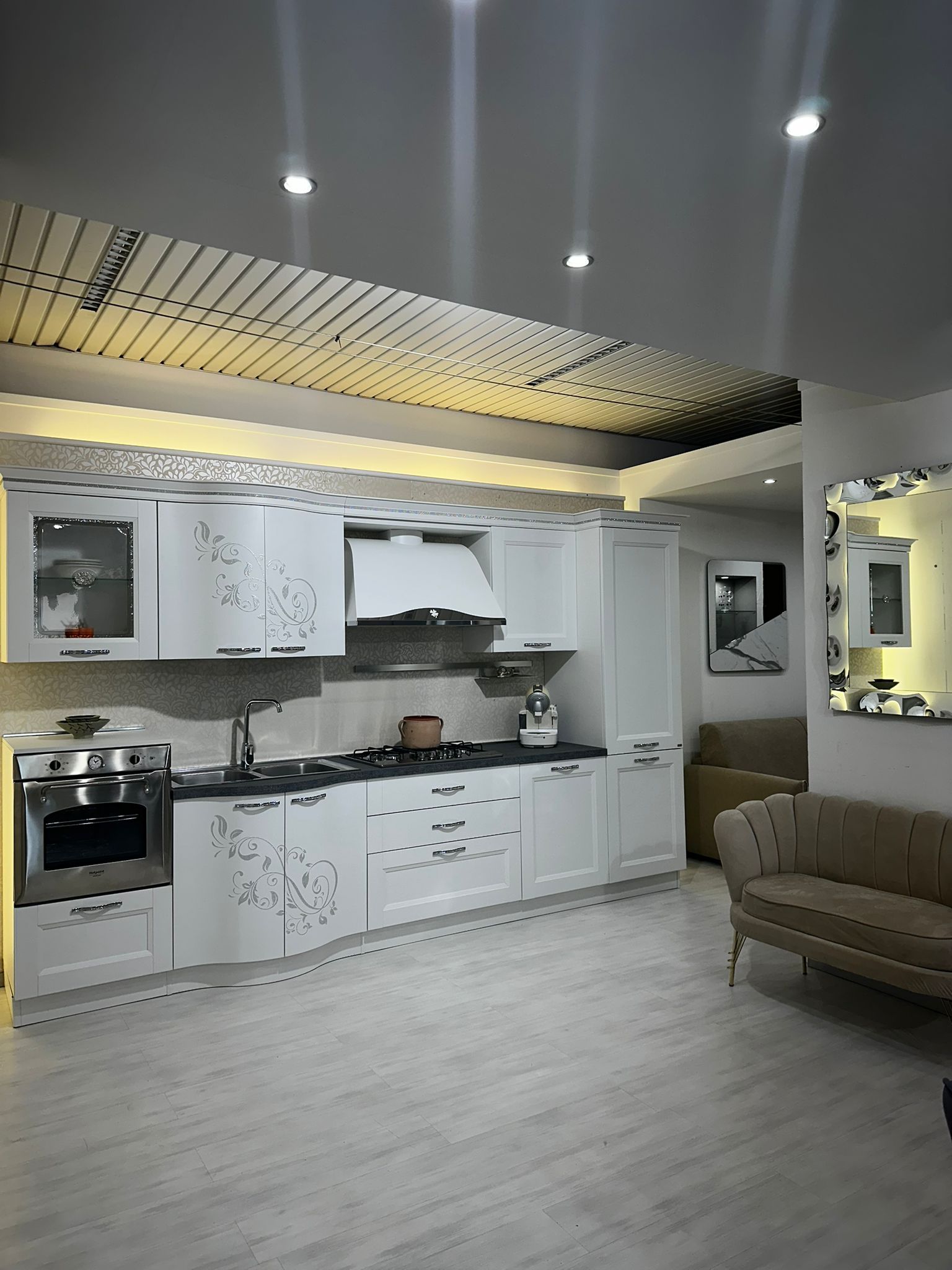 Cucina bianca con mobili moderni, fornelli e un divano sullo sfondo.