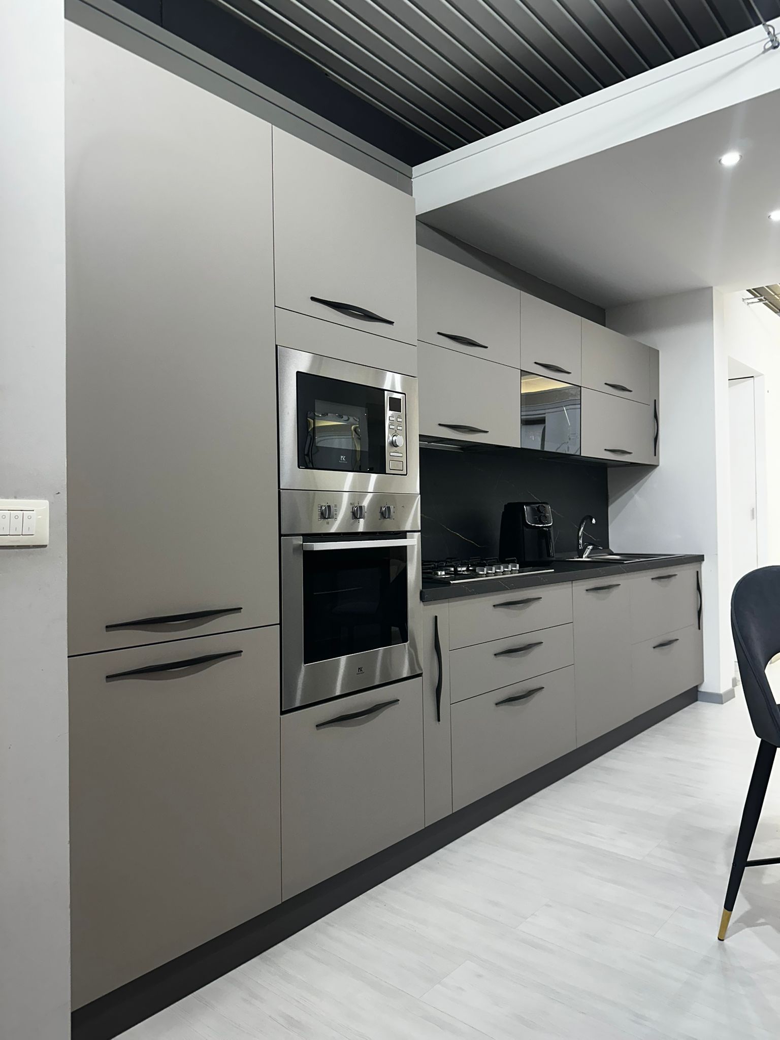 Moderna cucina grigia con elettrodomestici da incasso, ripiani scuri e mobili con maniglie nere.