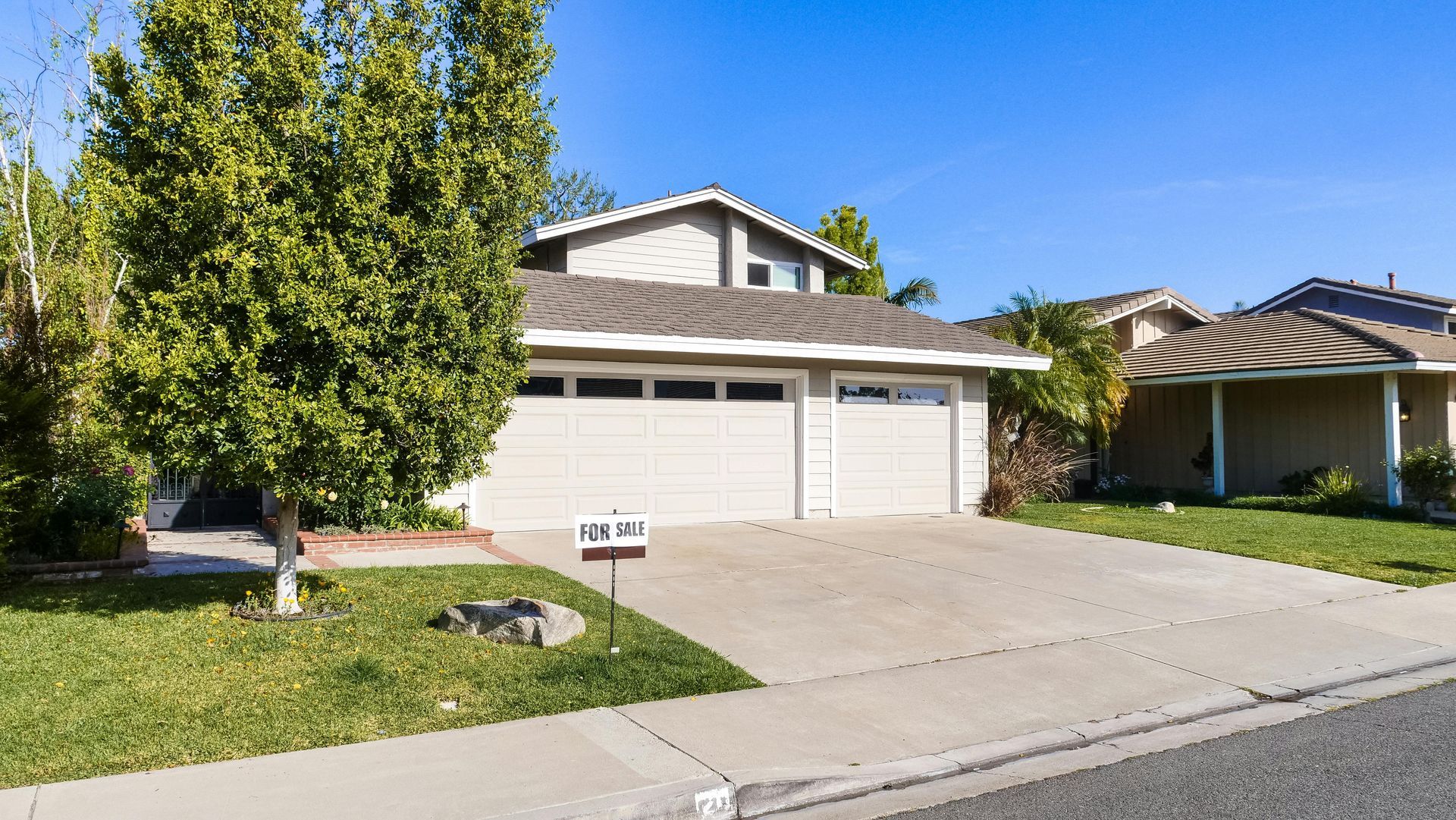 A two-story suburban house with a beige exterior, a two-car garage, and a manicured front lawn under a clear blue sky.
