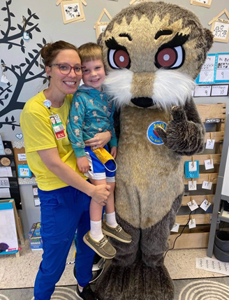 A woman is holding a child next to an otter mascot.