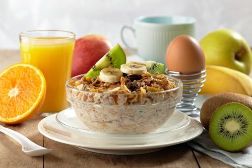 A bowl of cereal with bananas , kiwi , apples , eggs and orange juice on a wooden table.