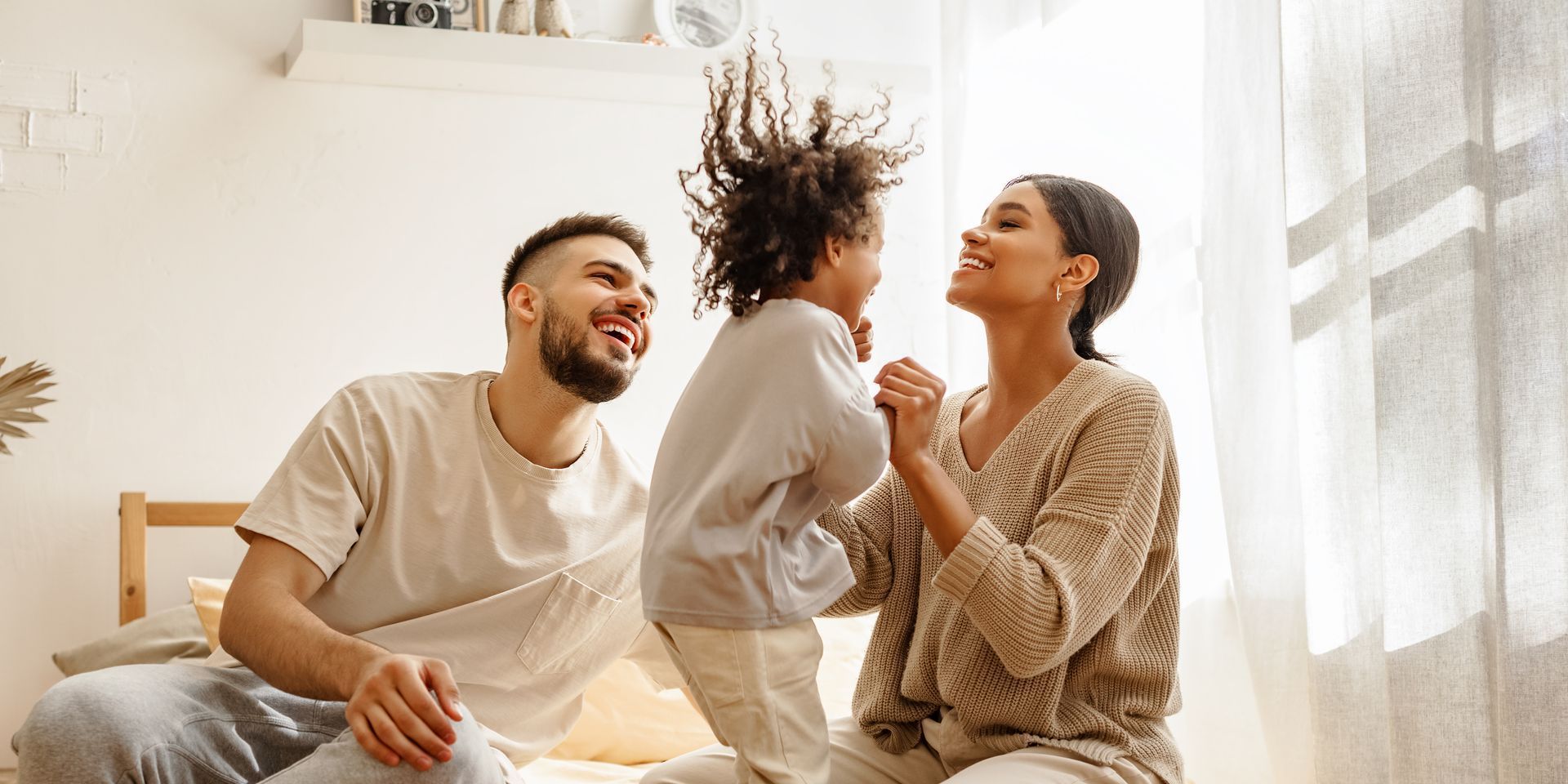 A family is sitting on a bed with a child jumping in the air.