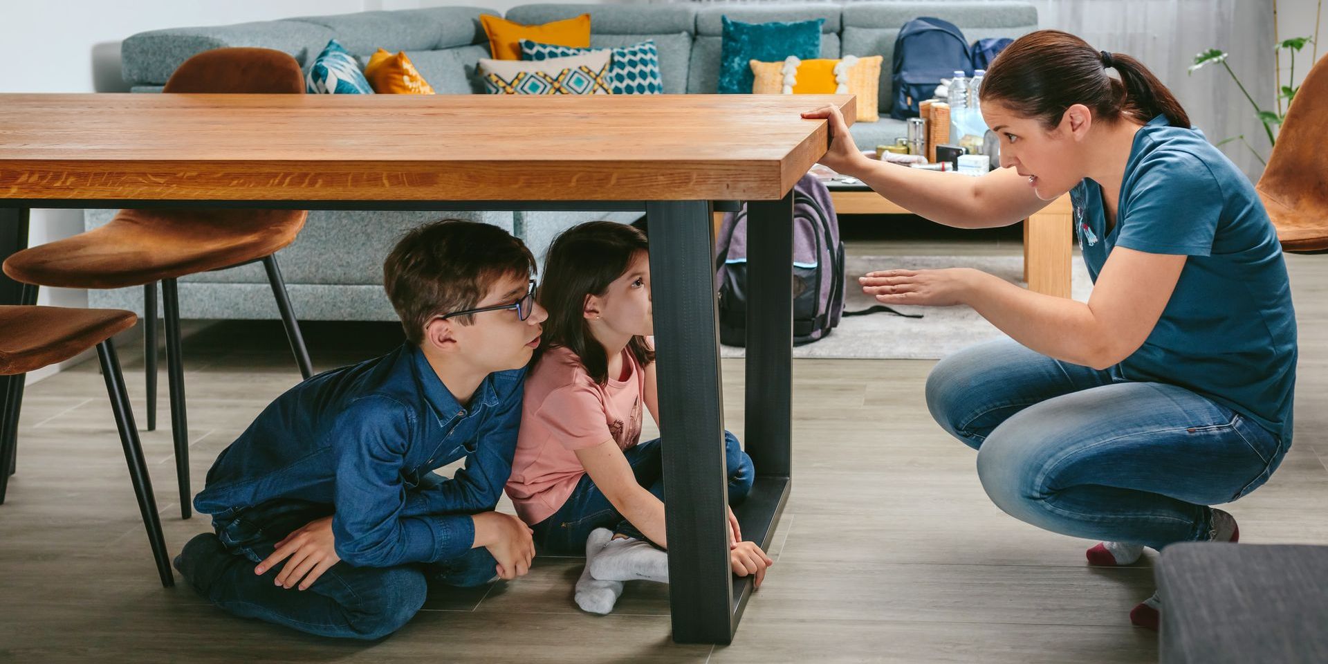 A woman and two children are hiding under a table in a living room.