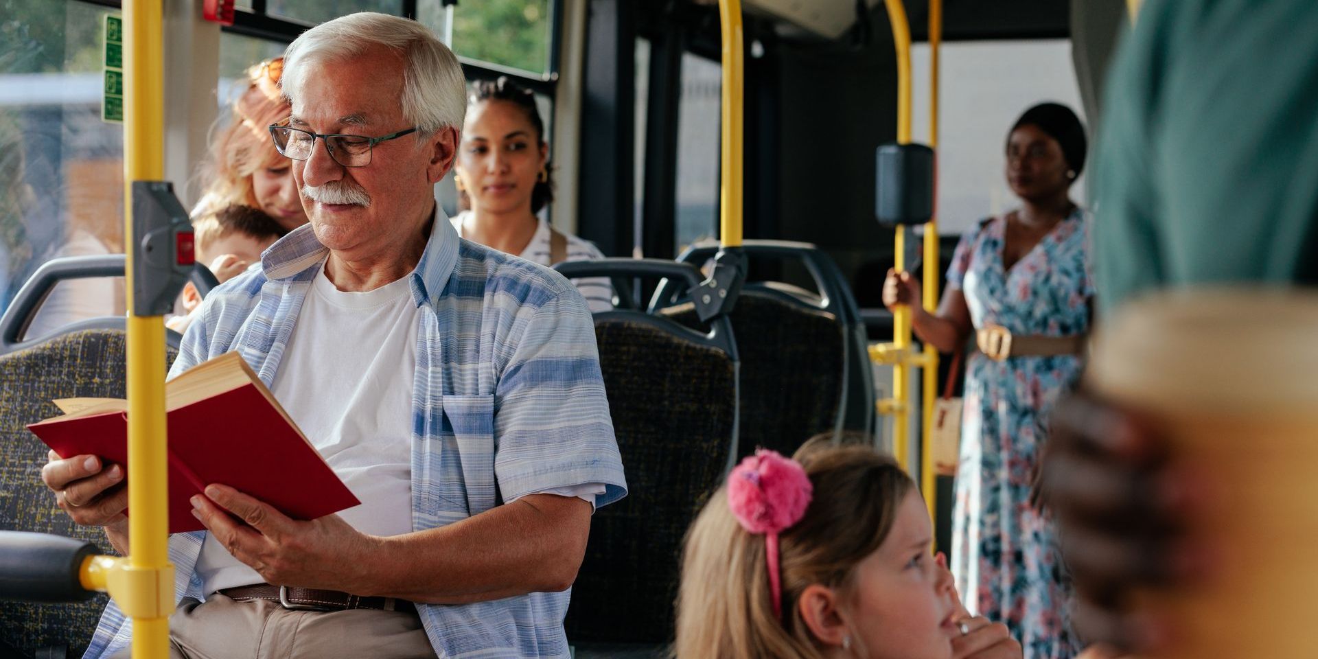 An elderly man is reading a book on a bus.