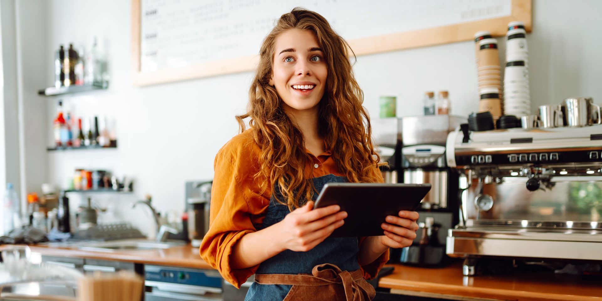 A woman in an apron is holding a tablet in a cafe.