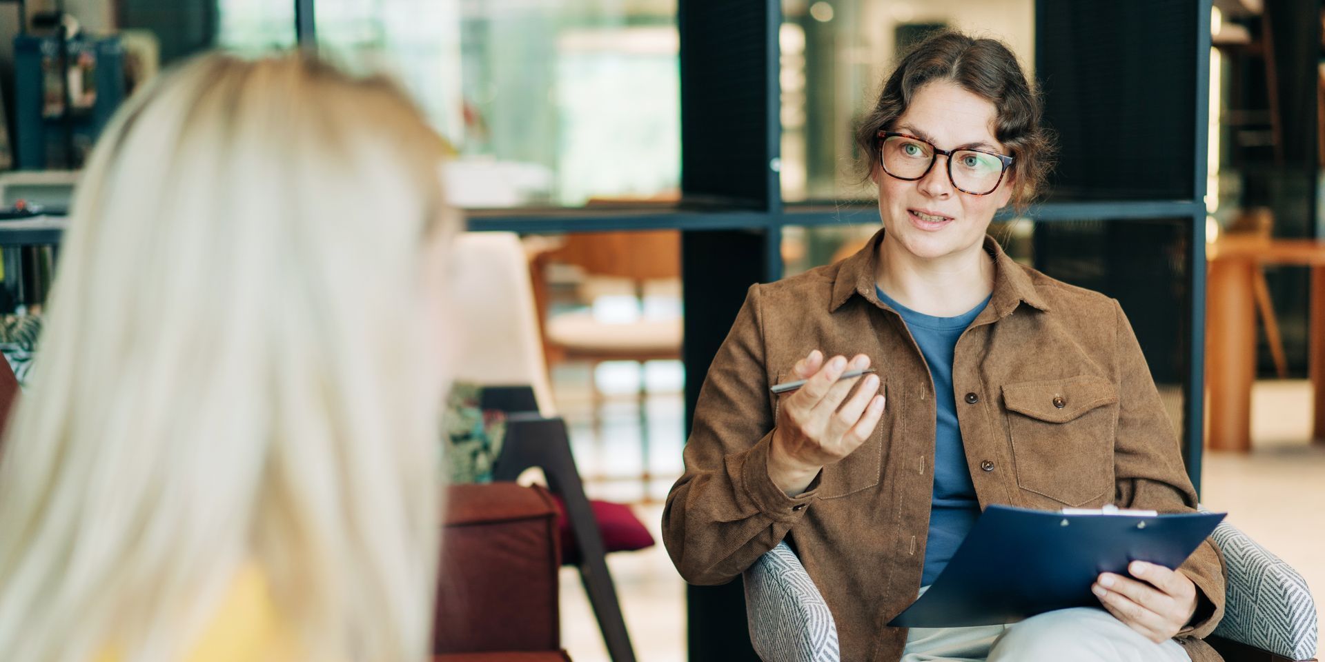 A man is sitting in a chair talking to a woman while holding a clipboard.