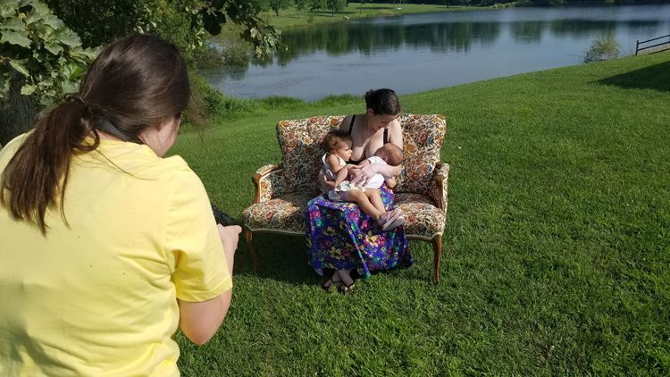 A woman getting her picture taken while breastfeeding two children on a couch outside