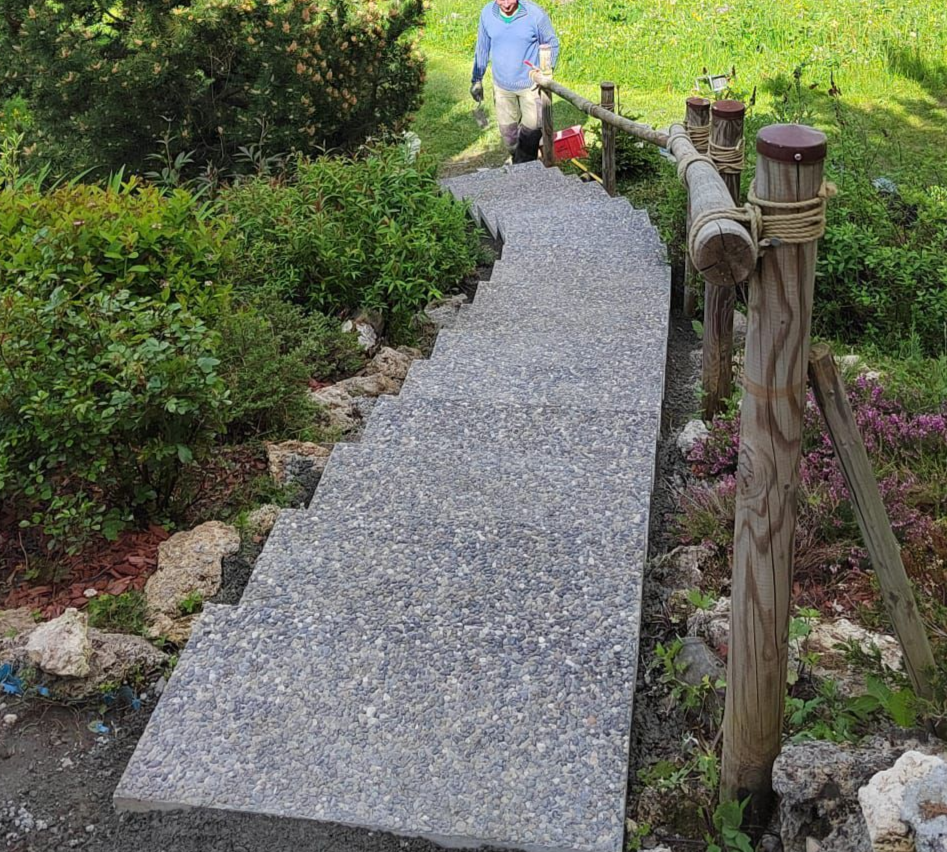 Allée de jardin incurvée en gravier avec des balustrades en bois, bordée de verdure, et une personne debout à l'extrémité.