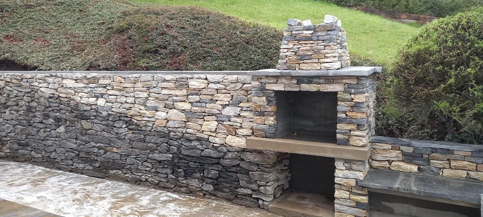 Cheminée extérieure en pierre intégrée à un mur de soutènement, avec des buissons et de l'herbe derrière.