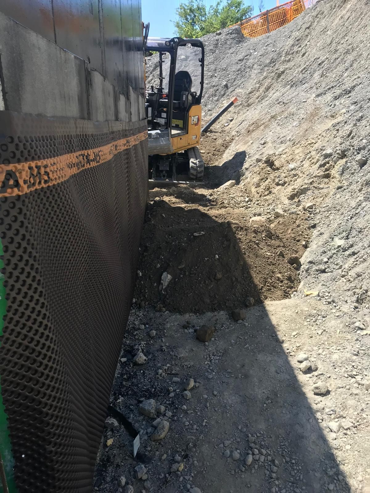 Une excavatrice travaille dans une étroite tranchée de terre, à côté d'un mur de soutènement et d'une pente de terrain abrupte.