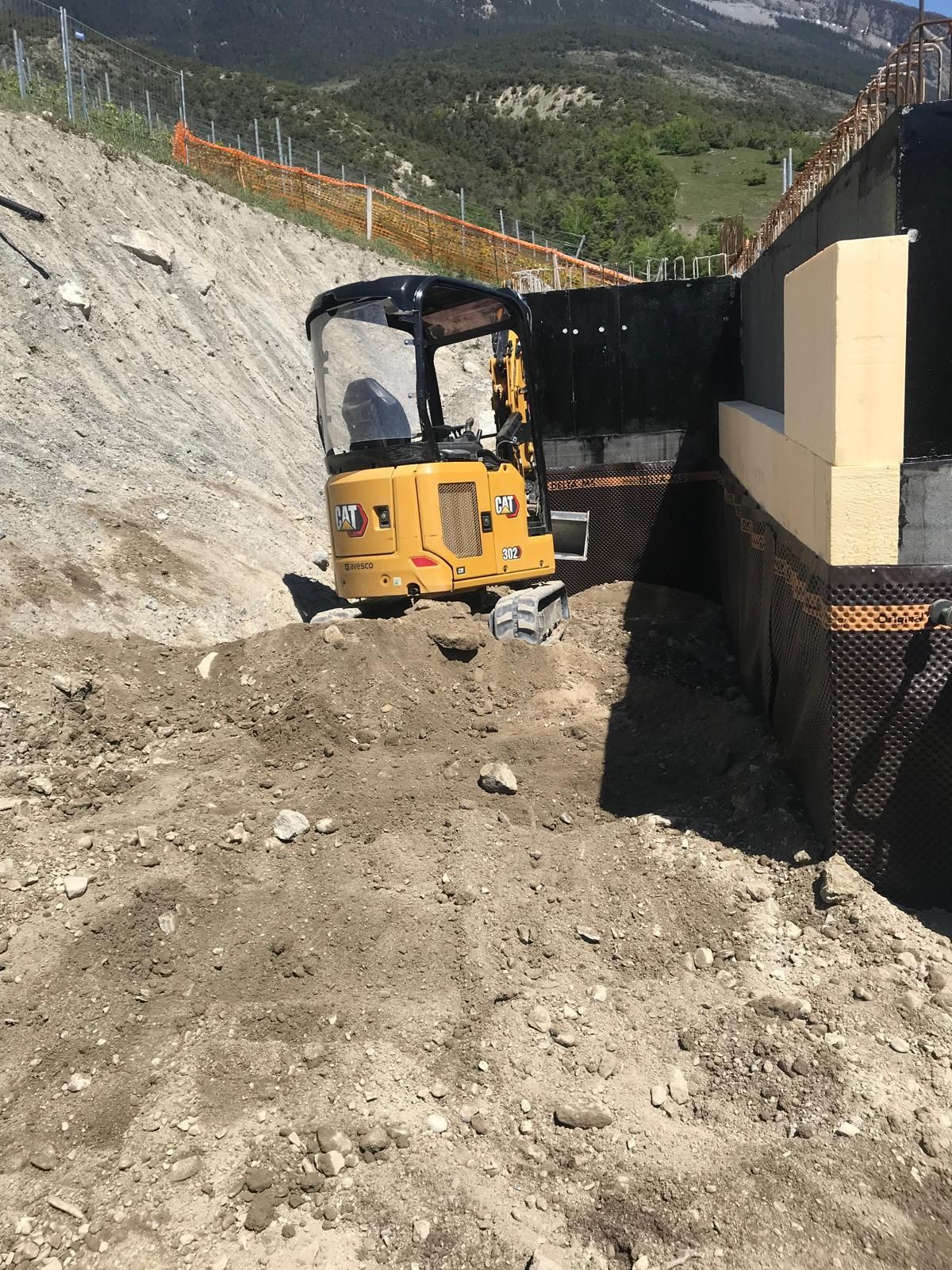 Mini-pelle jaune sur un talus de chantier rocailleux, à côté d'un mur de soutènement et d'une clôture de sécurité.